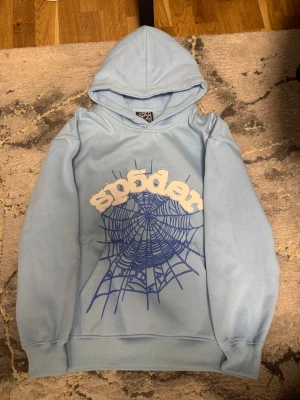 Ljusblå hoodie från SP5DER - Säljer en helt ny aldrig använd ljusblå hoodie från SP5DER med stor vit text och blått spindelnätsmönster på framsidan. Hoodien har huva och ribbade muddar vid ärmslut och nederkant. Perfekt för dig som gillar streetwear och unika prints.