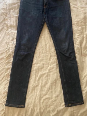 Mörkblå skinny jeans från Nudie - Säljer ett par mörkblå skinny jeans från Nudie med klassisk femficksdesign och snygga detaljer på bakfickorna. Jeansen har smal passform och är tillverkade i stretchigt denim för extra komfort.