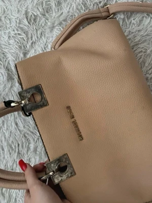 Beige axelremsväska från Michael Kors - Snygg beige axelremsväska från Michael Kors i strukturerat läder. Det finns tecken på slitage (se bilder).Väskan har två handtag och detaljer i metall med fyrkantiga fästen. Perfekt för dig som gillar stilrena och klassiska accessoarer.