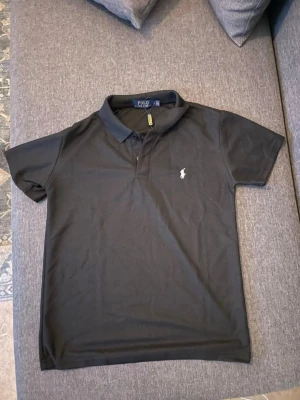  Polo Ralph Lauren pike - Klassisk svart pikétröja från Polo Ralph Lauren med kort ärm och vit broderad logga på bröstet. Tröjan har krage och knäppning framtill, perfekt för en stilren och enkel look.