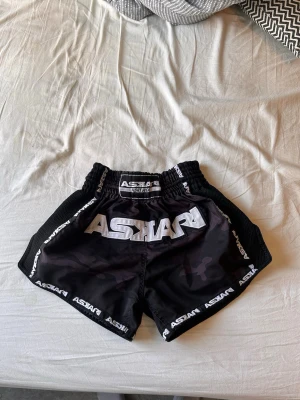 Svarta kamouflage shorts från Askari - Snygga svarta träningsshorts från Askari med diskret kamouflagemönster och vita logotyper längs kanterna. Shortsen har elastisk midja och meshpaneler på sidorna för extra ventilation. Perfekta för träning eller kampsport.