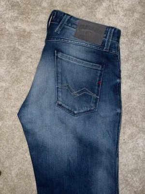Blå Replay jeans Anbass herr - Snygga blå jeans från Replay, modell Anbass. Jeansen har en klassisk femficksdesign, tydliga slitningar och Replay-logga på fickan. Tillverkade i stretchigt denim för skön passform. Perfekta till en avslappnad streetstil.