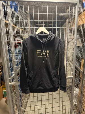 Svart hoodie från EA7 Emporio Armani - Snygg svart hoodie från EA7 Emporio Armani med stor logga i vitt på bröstet. Klassisk huva med dragsko och känguruficka framtill. Tillverkad i mjukt material som passar perfekt till en avslappnad stil.