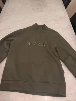 Olivgrön half zip sweatshirt från NICCE - Säljer en olivgrön sweatshirt från NICCE med halv dragkedja och broderad logga på bröstet. Tröjan har hög krage och är gjord i mjukt bomullsmaterial. Perfekt för en chill och stilren look.