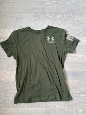 Grön Under Armour Freedom t-shirt - Snygg militärgrön t-shirt från Under Armour med trycket 'FREEDOM' och logga på bröstet. Amerikansk flagga på ärmen. Klassisk rund hals och korta ärmar. Tillverkad i mjuk bomull för skön känsla.