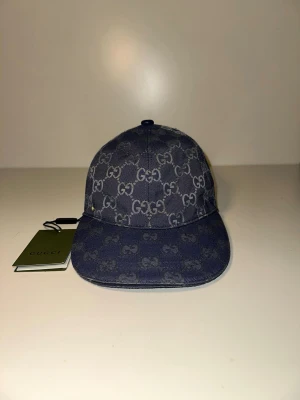 Blå GG-monogram keps från Gucci - Snygg blå keps från Gucci med klassiskt GG-monogram över hela kepsen. Tillverkad i denimliknande material och har böjd skärm. Märkets logga syns tydligt och kepsen har en modern, avslappnad look. Perfekt accessoar för dig som gillar exklusiv streetwear.