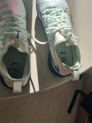 Nike Mercurial CR7 innefotbollsskor/gympaskor - Snygga Nike Mercurial CR7 Nike Zoom Vapor 16 Academy Mds Ic Sfotbollsskor i vitt med blå och gula detaljer. Skorna har låg profil, snörning och CR7-logga på innersulan. Tillverkade i syntetmaterial för bra grepp och känsla på planen. Perfekta för dig som vill sticka ut på fotbollsplanen.