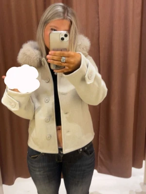 beige kappa från gina young 14+ - Snygg kort kappa i ljusbeige med stor fluffig fuskpäls runt kragen. Kappan har knappar framtill, två fickor och breda ärmslut med dekorativa knappar. Perfekt för dig som vill ha en trendig och mysig look.