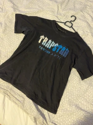 Trapstar svart t-shirt med tryck - Säljer en svart t-shirt från Trapstar med stort grafiskt tryck i vitt och blått på bröstet. Klassisk rund hals och korta ärmar. Perfekt för dig som gillar streetwear och vill ha en statement tee i garderoben.