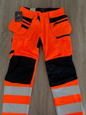 Varselbyxor från Björnkläder  - Säljer ett par orangea och svarta varselbyxor från björnkläder med flera praktiska fickor och reflexdetaljer. Byxorna har förstärkta partier på knän och ben samt breda reflexband för extra synlighet. Perfekta för dig som vill sticka ut på jobbet!