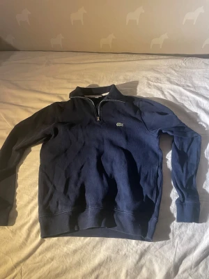 Mörkblå half-zip tröja Lacoste - Mörkblå långärmad tröja från Lacoste med klassisk half-zip och ståkrage. Tröjan har det ikoniska krokodilmärket broderat på bröstet och ribbade muddar vid ärmslut och nederkant. Perfekt för en avslappnad men stilren look.