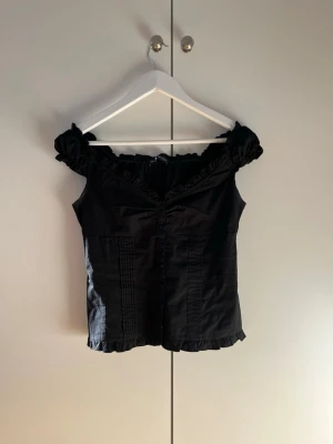Svart offshoulder blus med volang - Superfin populär svart offshoulder blus från Brandy Melville med volangdetaljer längs halsringningen och nederkanten. Blusen har knappar framtill och korta puffärmar. 