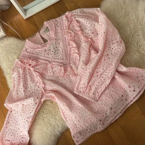 Rosa broderad blus med volanger - Söt ljusrosa blus med broderade blommor och hålmönster. Blusen har volangdetaljer vid bröstet och längs axlarna samt en hög krage med spetskant. Långärmad modell i tunn bomull, perfekt för en romantisk look.