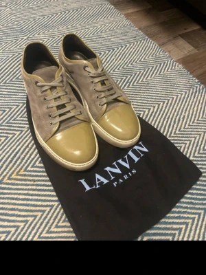 Lanvin sneakers i mocka och lack - Snygga sneakers från Lanvin i grå mocka med olivgröna lackdetaljer på tån och matchande snörning. Klassisk låg modell med vit sula och exklusiv känsla. Perfekta för dig som vill ha något unikt och stilrent från ett high-end märke.