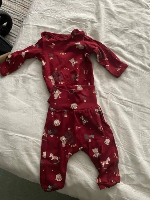 Rött babyset med nalle- och julmotiv - Sött tvådelat babyset i rött med långärmad body och byxor. Setet har gulliga tryck med nallar, trummor, gunghästar, presenter och julgranar. Mjukt och stretchigt material, perfekt för mysiga dagar. Passar perfekt för vintern och julen.
