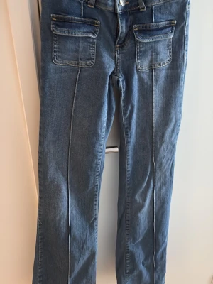 Blå jeans med fickor framtill - Säljer ett par blå bootcut jeans och markerade sömmar framtill. Jeansen har två stora fickor med lock på framsidan och låg midja. Perfekta för en avslappnad och trendig stil.