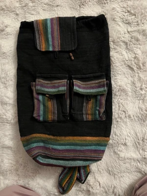 Svart ryggsäck med färgglada ränder - Unik svart ryggsäck i tyg med färgglada ränder i grönt, lila, gult och orange. Två framfickor med lock och träknappar samt ett större lock upptill. Justerbara axelband i samma randiga tyg. Perfekt för dig som gillar boho-stil och vill sticka ut.