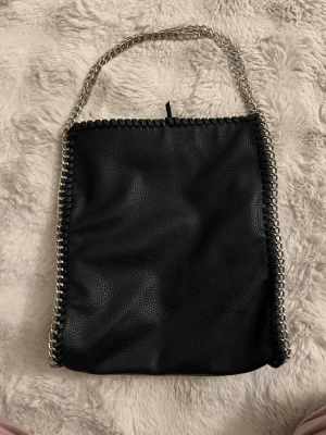 Svart handväska med kedjedetaljer ginatricot - Snygg svart handväska i fejkläder med silverfärgad kedja som axelrem och detaljer längs kanterna. Väskan har en mjuk, fyrkantig form och innerfack med dragkedja. Perfekt för dig som gillar en edgy och trendig look.