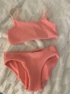 Rosa ribbad bikini med cropptopp - Trendig rosa bikini med ribbad struktur och cropptopp. Överdelen har smala axelband och underdelen är klassiskt skuren. Perfekt för strandhäng eller poolparty. Materialet känns mjukt och följsamt mot huden.