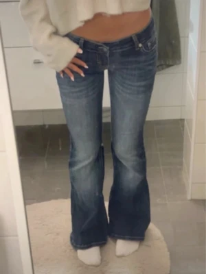 Blå bootcut jeans med låg midja - Säljer ett par blå bootcut jeans med låg midja och klassiska femficksdetaljer. Jeansen har en snygg tvätt med lätt slitning och är tillverkade i denim. Perfekta för dig som gillar en avslappnad och trendig look.