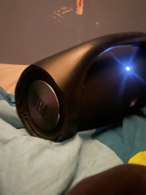 JBL Boombox svart trådlös högtalare - Stor och kraftfull JBL Boombox i svart med robust handtag och rundad form. Högtalaren har JBL-logga på sidan och är tillverkad i slitstark plast och tygmaterial. Perfekt för att spela musik trådlöst via Bluetooth och har tydliga basreflexer på sidorna.