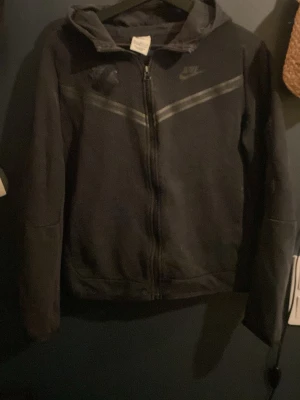 Svart Nike hoodie med dragkedja XL - Säljer en svart hoodie från Nike i storlek XL. Hoodien har dragkedja framtill, huva och en diskret Nike-logga på bröstet. Snygg detalj med en diagonal grå linje över bröstet. Perfekt för dig som gillar sportig och enkel stil.