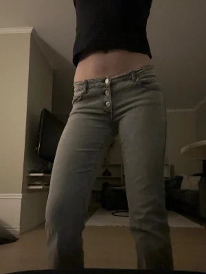 Gråa bootcut jeans med knappar - Säljer ett par gråa jeans med utsvängda ben och låg midja. Jeansen har tre synliga silverfärgade knappar framtill. Perfekta för en avslappnad och trendig look.