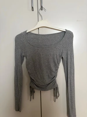 Grå ribbad långärmad topp med snörning - Snygg grå ribbad topp med långa ärmar och snörning i sidorna som ger en croppad look. Perfekt till jeans eller kjol för en trendig stil. Mjuk och stretchig, passar dig som gillar enkel men cool design.