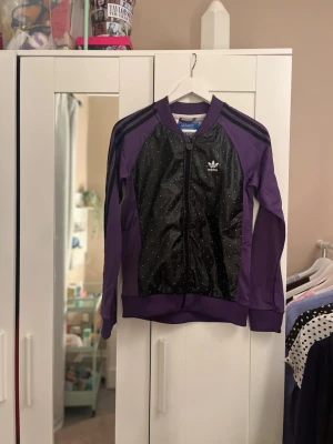 Lila Adidas ziptröja med svarta detaljer - Säljer en lila ziptröja från Adidas med svarta paneler framtill som har små vita prickar. Tröjan har klassiska Adidas-ränder på axlarna, ribbade muddar och dragkedja hela vägen. Perfekt för en sportig och avslappnad stil.