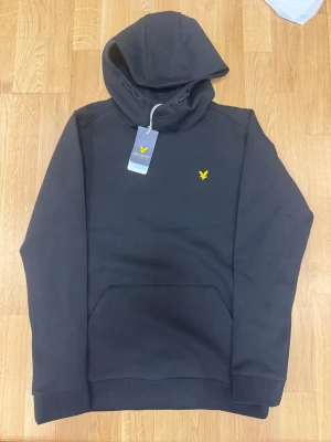 Svart hoodie från Lyle & Scott - Helt ny med prisa lappen Svart hoodie från Lyle & Scott med klassisk gul logga på bröstet. Hoodien har en stor huva med dragsko och en rymlig magficka. Tillverkad i mjukt material som känns skönt mot huden. Perfekt för en avslappnad och stilren look.