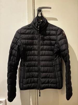 Svart dunjacka från Parajumpers - Snygg svart dunjacka från Parajumpers med quiltad design och hög krage. Jackan har dragkedja framtill, två sidofickor med dragkedja och en patch med logga på ärmen. Perfekt för kyliga vår/höst dagar och riktigt lätt att bära. Jackan är i bra skick men har ett litet hål på backsidan samt några sömmar på insidan som gått uppåt. (Påverkar inte användningen)