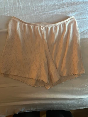 Ljusa pyjamasshorts från Cubus - Säljer ett par ljusa pyjamasshorts från Cubus i mjukt, ribbat material med diskreta ränder och små hålmönster. Shortsen har en liten rosett i midjan och vågig kant nertill för en söt look. Perfekta för mysiga kvällar hemma.