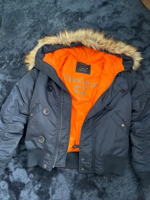 Svart bomberjacka från Crocker - Stilren svart bomberjacka från Crocker med orange foder och avtagbar huva med fuskpäls. Jackan har ribbade muddar, dragkedja och knappar framtill samt flera fickor. Perfekt för dig som vill ha en klassisk men ändå cool look.