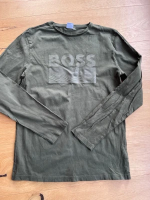 Mörkgrön långärmad BOSS t-shirt - Säljer en mörkgrön långärmad t-shirt från BOSS med snyggt tryck på bröstet. T-shirten har en klassisk rund halsringning och är tillverkad i mjuk bomull som känns skön mot huden. Perfekt för dig som gillar stilrena plagg med diskret logga.