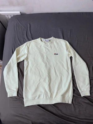Ljusgul sweatshirt från FILA - Säljer en ljusgul sweatshirt från FILA med liten broderad logga på bröstet. Tröjan har rund halsringning, långa ärmar och ribbade muddar vid ärmslut och nederkant. Perfekt för en avslappnad och sportig stil.