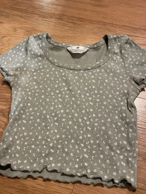 Grön blommig croppad topp H&M - Söt grön croppad topp från H&M med små vita blommor och ribbad struktur. Toppen har korta ärmar, rund halsringning och vågig kant nertill. Perfekt för dig som gillar en avslappnad men trendig stil. Storlek 134-140