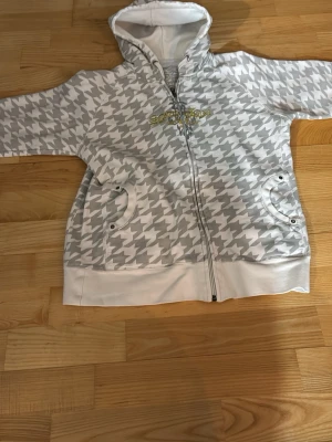 Vit och grå hoodie med dragkedja - Säljer en vit och ljusgrå hoodie med dragkedja och stort hundtandsmönster. Tröjan har huva, två fickor framtill och ribbade muddar. Detaljer i silver och guldtext på bröstet. Perfekt för en chill och avslappnad stil.