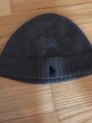 Mörkgrå stickad mössa från Polo Ralph Lauren - Säljer en mörkgrå stickad mössa från Polo Ralph Lauren med klassisk ribbad kant och broderad marinblå logga framtill. Mössan är mjuk och skön, perfekt för kalla dagar och har en enkel, stilren design som passar till det mesta.