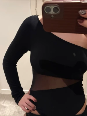 Svart mesh-detaljerad body - Säljer en svart body med en ärm och mesh-detaljer över magen. Snygg asymmetrisk design med en axel bar och lång ärm. Materialet är stretchigt 