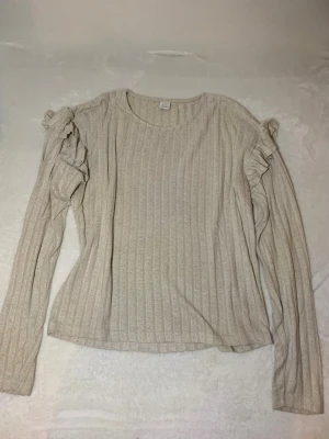 Beige ribbstickad tröja med volanger - Snygg beige ribbstickad tröja med långa ärmar och volangdetaljer längs axlarna. Tröjan har rund halsringning och är tillverkad i ett mjukt, stretchigt material som sitter skönt på kroppen. Perfekt för dig som gillar en enkel men trendig look.