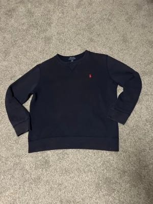Mörkblå sweatshirt från Polo Ralph Lauren - Klassisk mörkblå sweatshirt från Polo Ralph Lauren med rund halsringning och den ikoniska röda logotypen broderad på bröstet. Tröjan har ribbade muddar vid ärmslut och nederkant, perfekt för en clean och stilren look.