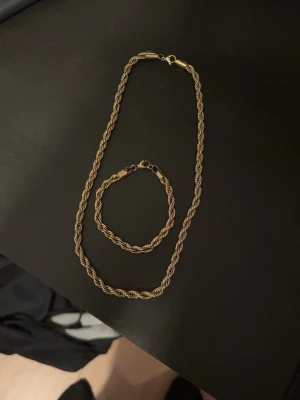 Guldfärgat tvinnat halsband och armband - Säljer ett stilrent halsband i guldfärgad metall med tvinnad kedja och matchande armband. Kedjan har en snygg, vriden design som ger ett exklusivt intryck. Perfekt accessoar för att lyfta din outfit med en touch av guld.
