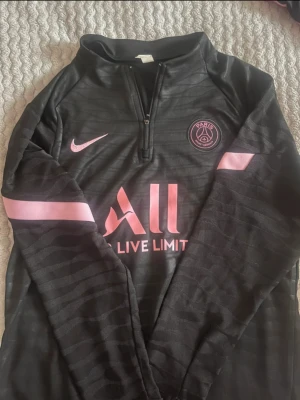 Svart PSG träningströja från Nike - Snygg svart långärmad träningströja från Nike med Paris Saint-Germain-logga och rosa detaljer. Halvlång dragkedja framtill och tryckta loggor på bröstet och ärmarna. Perfekt för fotbollsträning eller chill. Materialet känns mjukt och sportigt. Inte äkta, köpt på Vinted