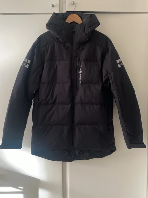 Svart dunjacka från Sail Racing - Säljer en svart dunjacka från Sail Racing med huva och dragkedja framtill. Jackan har logotyp på ärmen och bröstet samt justerbar huva med snörning. Perfekt för kalla vinterdagar tack vare det tjocka dunfodret och den stilrena designen.