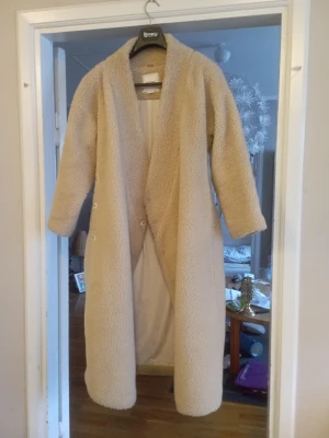 Beige lång teddyjacka med bälte - Mysig och lång teddyjacka i beige med bälte i midjan och långa ärmar. Jackan har en öppen design utan synliga knappar framtill och är fodrad på insidan för extra komfort. Perfekt för kalla dagar när du vill vara både varm och trendig. Är en oversized modell.