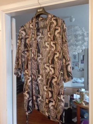 Svart kimonomorgonrock med hägerprint - Snygg svart morgonrock i kimonomodell från Linnea, täckt av ett coolt mönster med vita hägrar och gröna inslag. Lätt och luftig med trekvartsärmar och knytskärp i midjan. Perfekt för chill dagar hemma eller när du vill känna dig extra stylish.