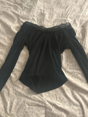 Svart topp med spetskant - Trendig svart offshoulder topp med lång ärm och snygg spetskant längs urringningen. Toppen har en smickrande passform och är perfekt för dig som vill ha en stilren men ändå detaljrik look. Materialet känns mjukt och följsamt. 