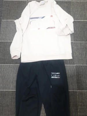 Beige och svart loungewear-set Calvin - Mysigt loungewear-set från Calvin med en beige sweatshirt med färgglada detaljer och logga framtill samt matchande svarta byxor med logga på benet. Tröjan har rund hals och muddar vid ärmslut och midja. Perfekt för chill dagar hemma.