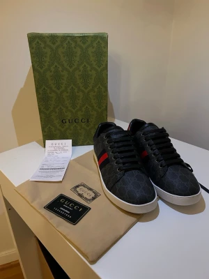 Gucci GG Supreme sneakers svart - Snygga Gucci sneakers i svart GG Supreme canvas med klassiska röda och gröna ränder på sidan. Skorna har vita sulor och metallic-detaljer i rött och blått på hälen. Perfekt för dig som vill ha en lyxig och trendig look.