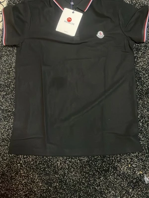 Svart pikétröja från Moncler - Klassisk svart pikétröja från Moncler med korta ärmar och diskret logga på bröstet. Kragen har tunna ränder i rött och vitt för en sportig touch. Perfekt till jeans eller shorts för en clean look.